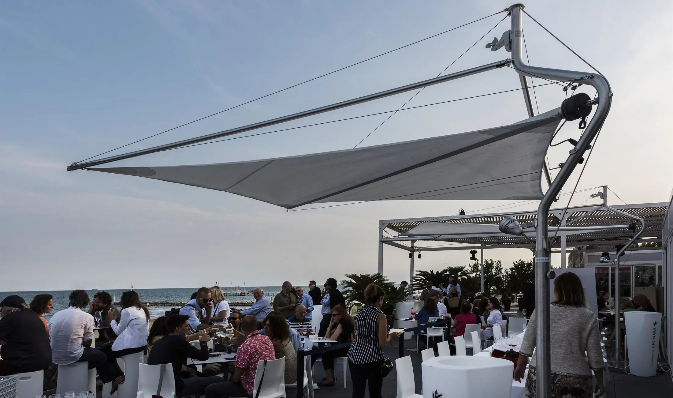 Mehrere Corradi Defense Sonnensegel schützen Sitzgelegenheiten eines Restaurants am Meer vor zu viel Sonneneinstrahlung.