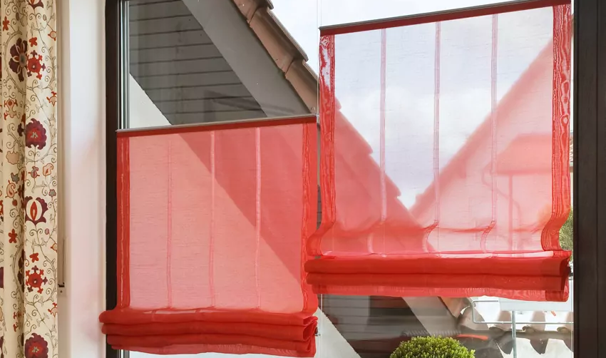 Rote Raffrollos mit transparentem Stoff setzen einen dezenten Farblichen Akzent und schirmen zu starke Sonne ab.