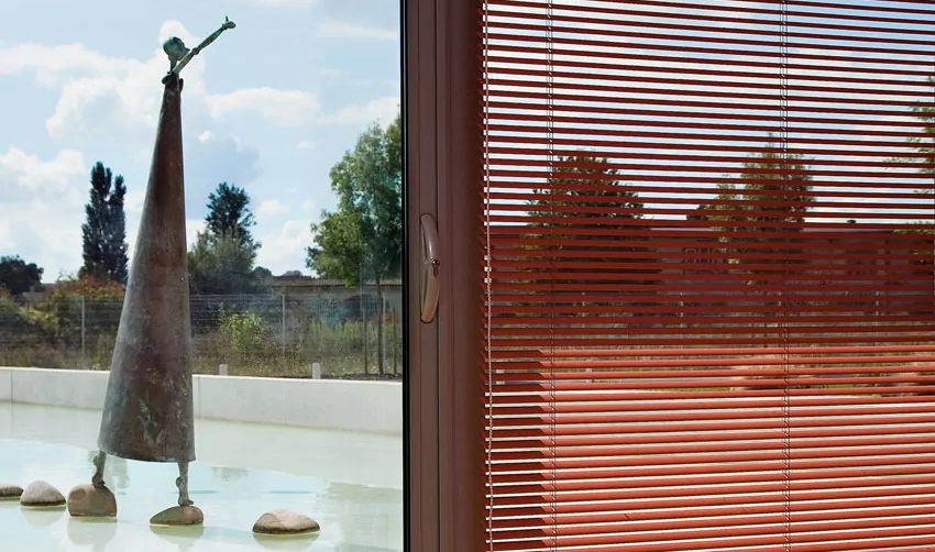Eine rötliche horizontale Jalousie von MHZ mit schmalen Lamellen reguliert den Lichteinfall an einem Fenster vor einem Wasserbecken mit einer großen Statue.