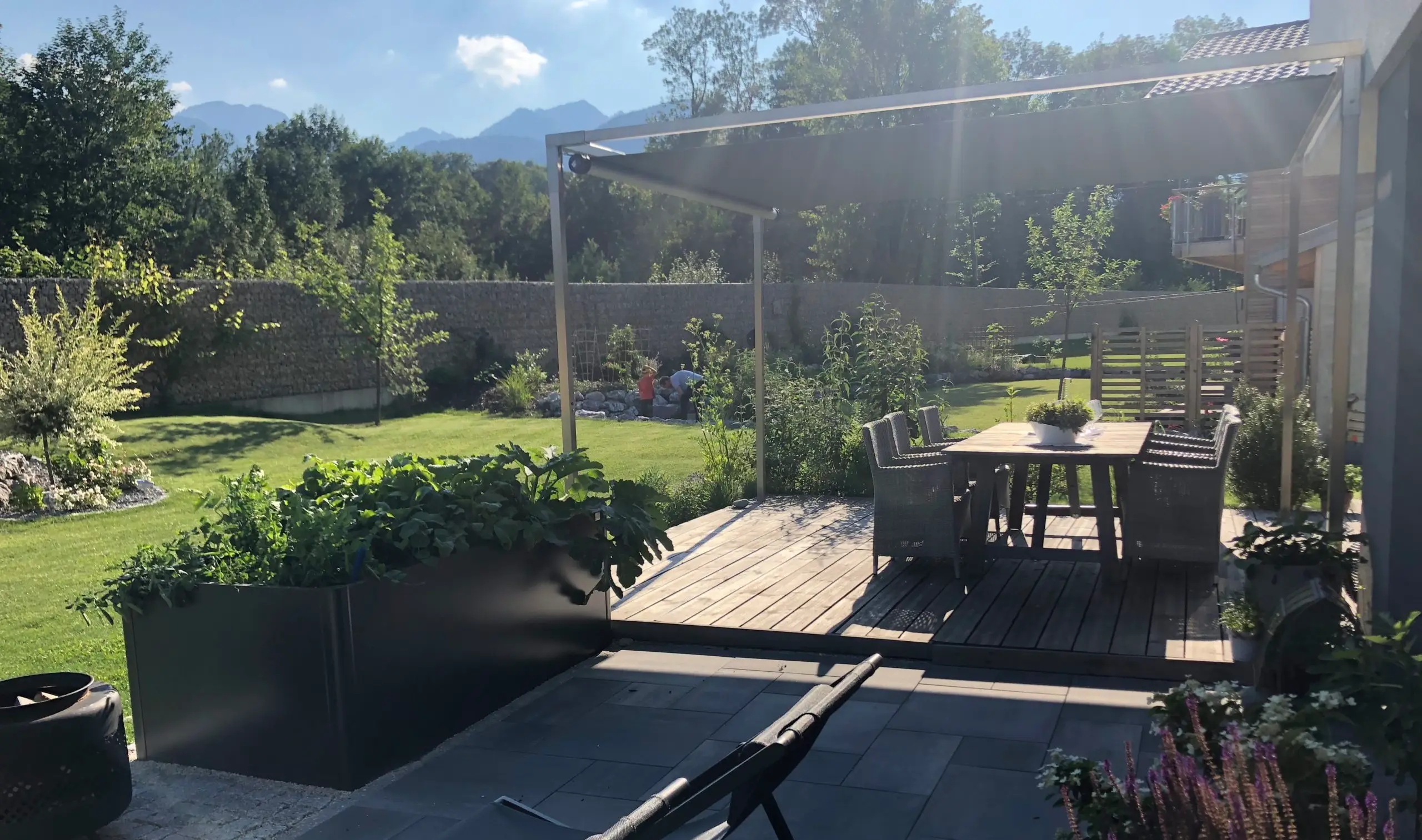 Ein Shadesign Shade CUBE ist über ein hölzernes Sonnendeck mit einem Sitzbereich im Garten gespannt.