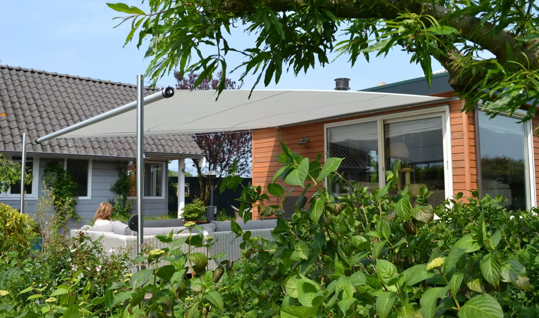 An einem hölzernen Bungalow beschattet das Sonnensegel Shadesign Shade INOX einen Outdoor-Sitzbereich, in dem eine Frau sitzt.