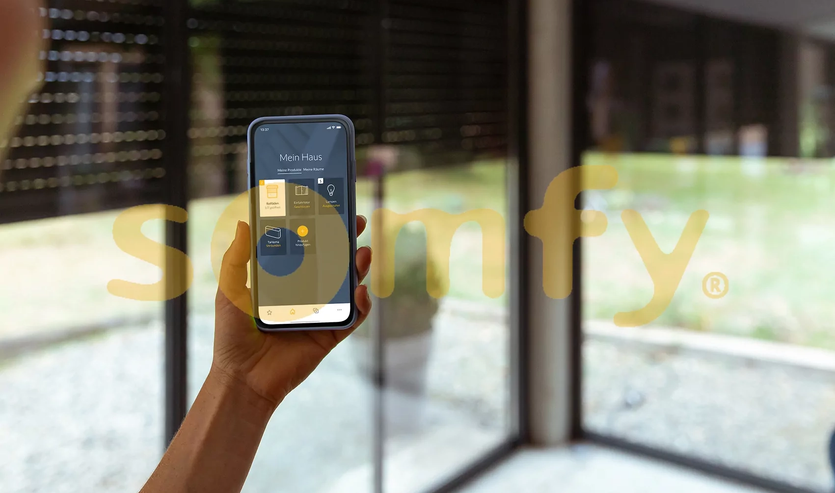 Die Oberfläche der Somfy Smartphone App vor Fenstern mit Rolläden hinter dem Somfy Logo.