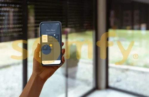 Somfy App auf einem Smartphone vor einem Fenster mit Rolladen und hinter dem Somfy Logo.
