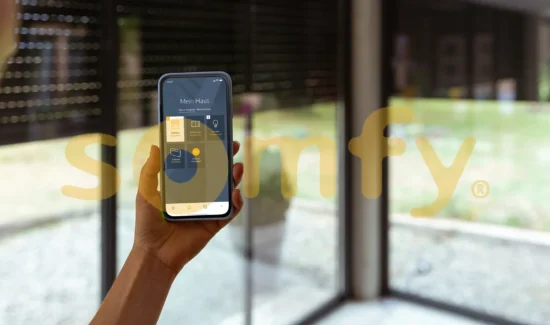 Somfy App auf einem Smartphone vor einem Fenster mit Rolladen und hinter dem Somfy Logo.