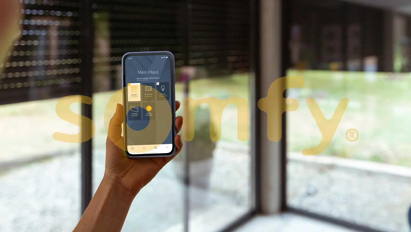 Somfy App auf einem Smartphone vor einem Fenster mit Rolladen und hinter dem Somfy Logo.