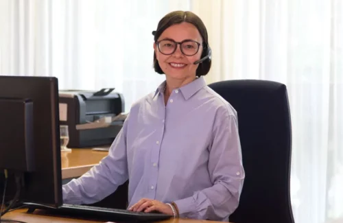 Frau mit Headset sitzt an einem Schreibtisch und lächelt in die Kamera.