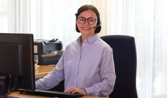 Frau mit Headset sitzt an einem Schreibtisch und lächelt in die Kamera.